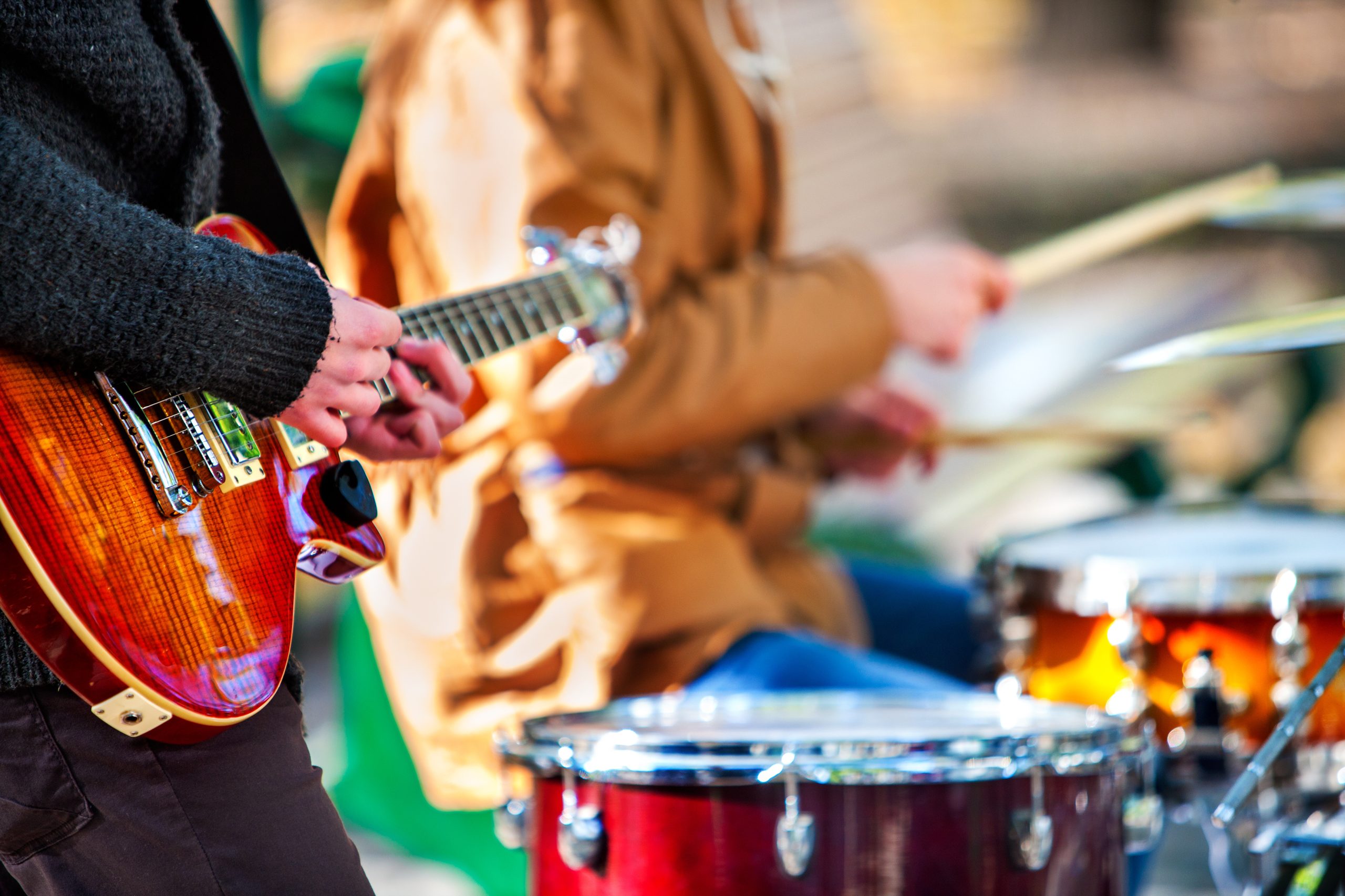 L'image présente une scène musicale en extérieur, mettant en avant deux musiciens en train de jouer de leurs instruments. À l'avant-plan, une personne vêtue d'un pull en laine épaisse de couleur foncée joue d'une guitare électrique au corps verni aux teintes rougeoyantes et aux reflets boisés. Ses doigts sont positionnés sur le manche de l'instrument, appuyant sur les cordes pour former des accords ou des notes individuelles. En arrière-plan, légèrement flou, un autre musicien joue de la batterie. Il porte une veste beige ou marron clair et tient une paire de baguettes, frappant les fûts et cymbales. Les éléments de la batterie visibles incluent une caisse claire rouge brillante et des cymbales montées sur des supports métalliques. L'arrière-plan est flou, mais laisse entrevoir un cadre extérieur avec une lumière naturelle vive, suggérant une performance en plein air. L'image met en évidence la gestuelle et la synchronisation des musiciens, capturant l'énergie d'une performance musicale.